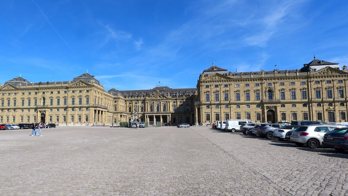 Panoramaaufnahme der Würzburger Residenz von vorne. Der weitläufige Residenzplatz führt auf die barocke Fassade des Schlosses zu, das mit seinen drei Flügeln den Ehrenhof umschließt. In der Mitte des Platzes ist der kunstvolle Frankoniabrunnen zu sehen, während die Sandsteinfassade im warmen Licht erstrahlt.